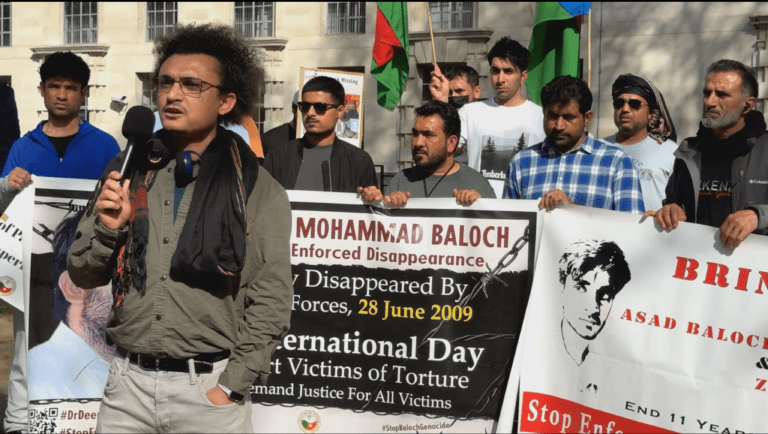Baloch political activist Aomar Karim delivering a speech at the Baloch National Movement UK Chapter protest outside 10 Downing Street, London, on 8 April 2026, with activists holding banners demanding justice for enforced disappearance victims including Mohammad Baloch and Asad Baloch.
