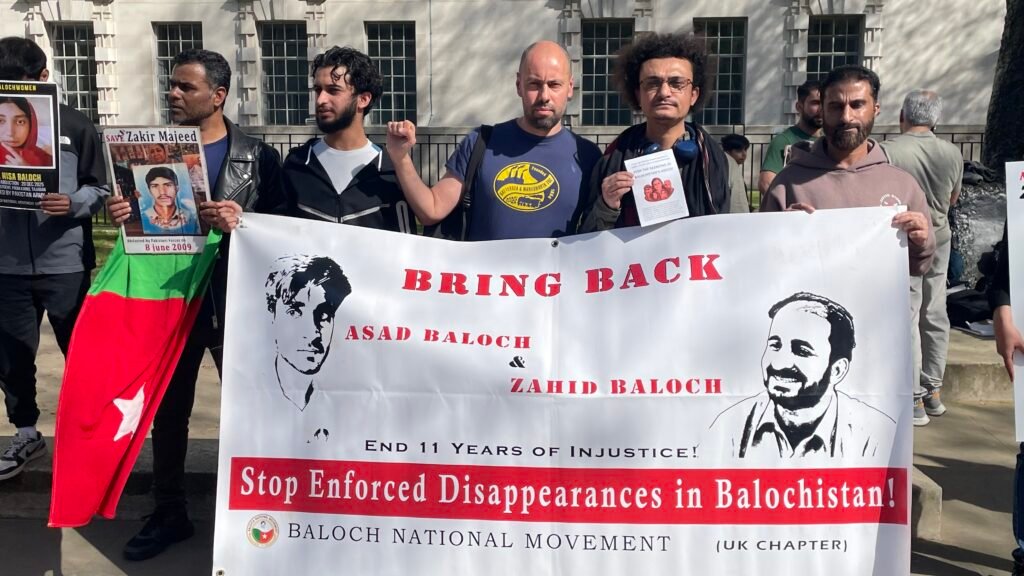 Baloch political activist Aomar Karim (right) and trade union activist Sacha Ismail (centre, raised fist) stand with fellow Baloch activists outside 10 Downing Street, London, holding a banner demanding the return of enforced disappearance victims Asad Baloch and Zahid Baloch, during the Baloch National Movement UK Chapter protest on 8 April 2026.